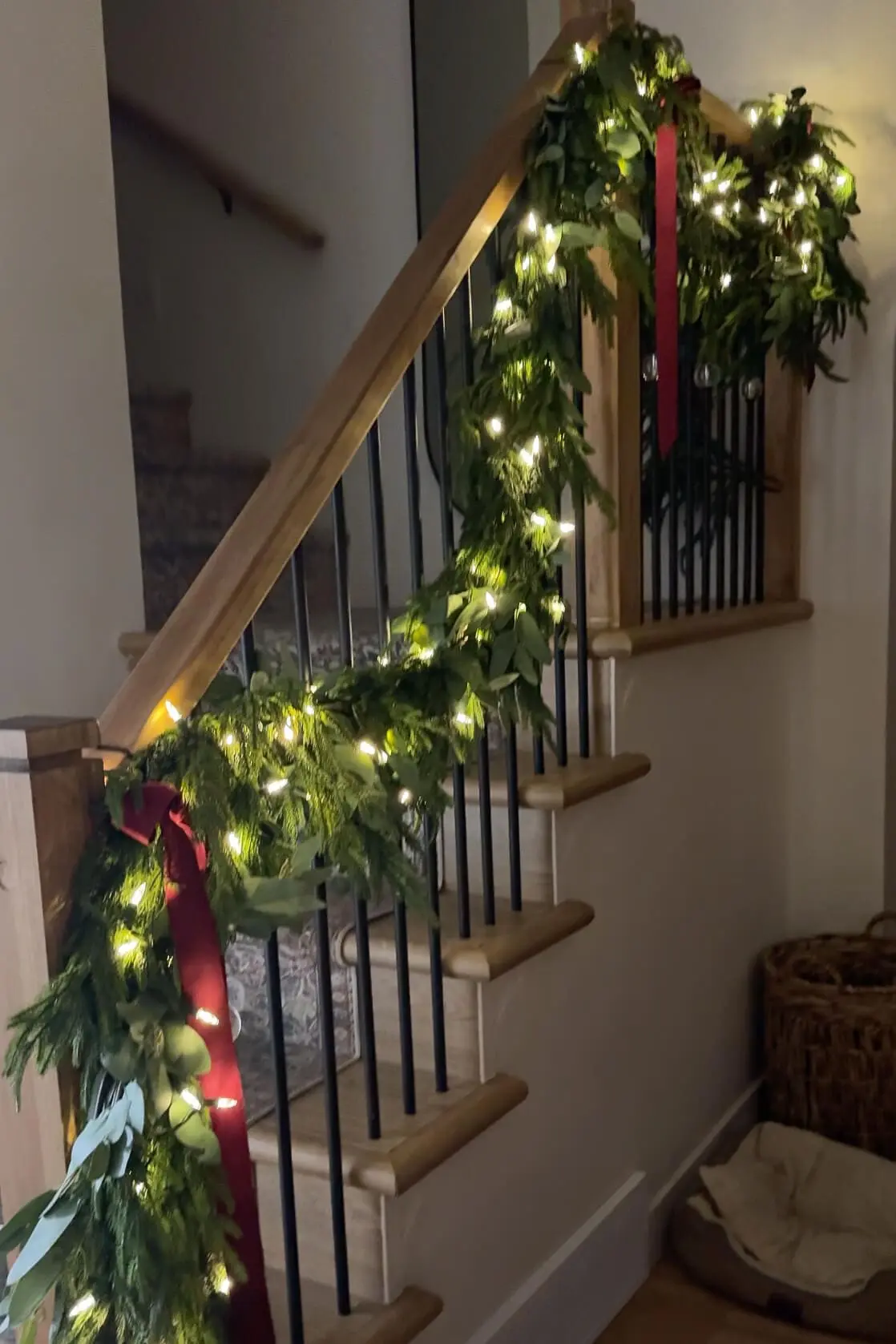 A warmly lit staircase adorned with a lush green garland featuring twinkling fairy lights and rich red velvet ribbon accents. The garland cascades along the wooden handrail, adding a festive touch to the entryway.
